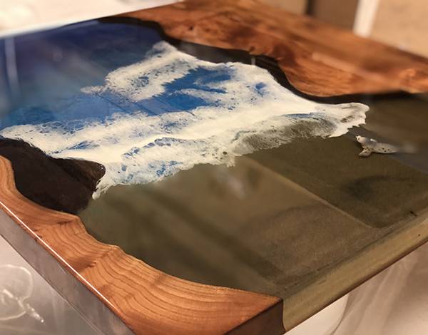Et nærbilde av et elvebord av tre med et harpiksinnlegg. Epoksydesignet skildrer en strandscene med blå, hvite og gjennomsiktige bølger som slår mot en sandstrand. Treet har en rik, naturlig åre og varme toner, som komplementerer kystestetikken til harpikskunstverket laget med Glassklar Epoxy - CCR Epoxy Entropy SuperSap.