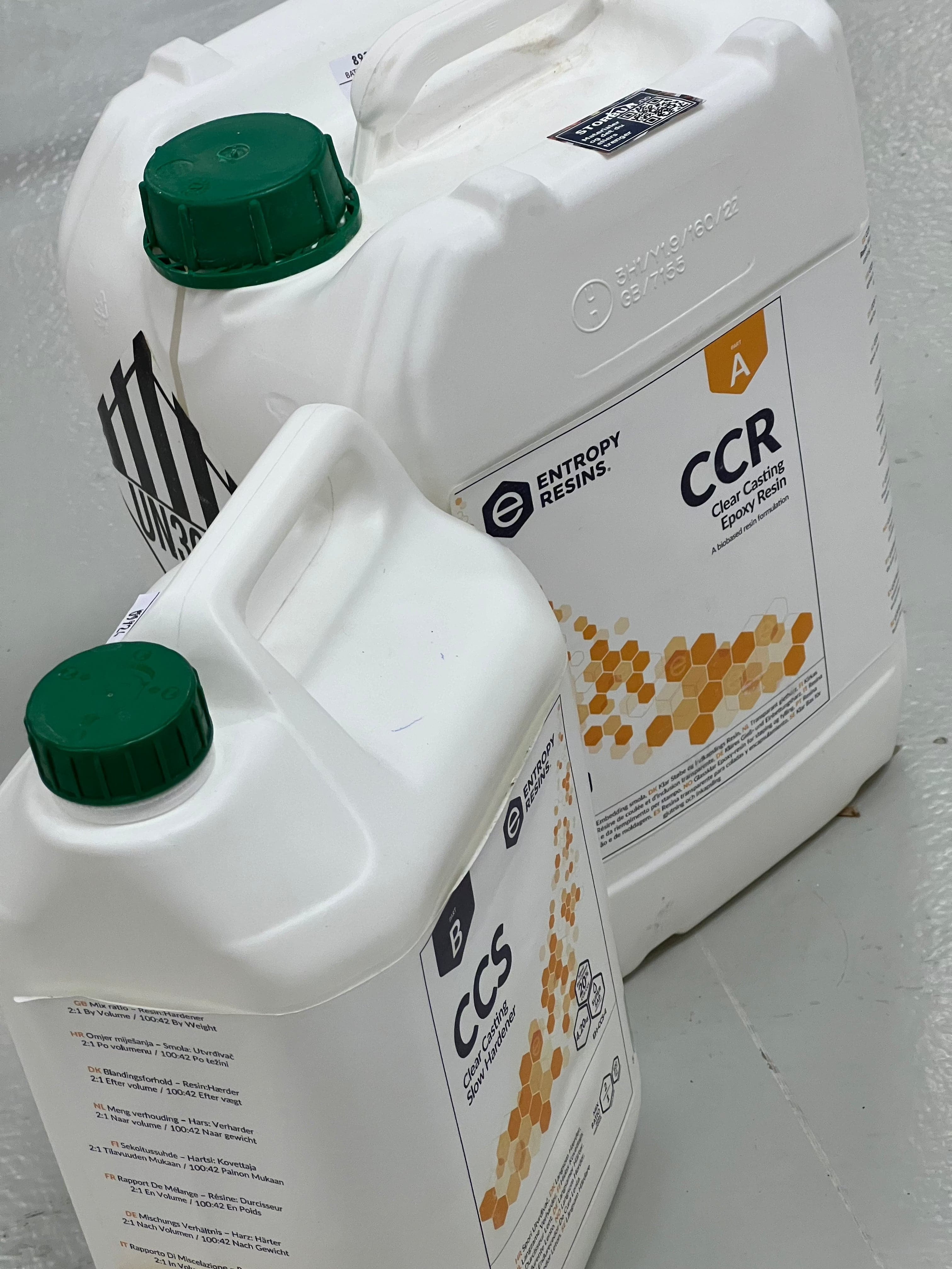 To hvite plastbeholdere med Glassklar Epoxy - CCR Epoxy Entropy SuperSap står på gulvet. Den større beholderen er merket "CCR SuperSap Clear Casting Epoxy Resin" med en oransje honeycomb-design, ideell for å lage et fantastisk elvebord. Den mindre beholderen, som er utformet på samme måte, er merket "CCS Clear Casting Epoxy Hardener." Begge har grønne capser og detaljert tekst.