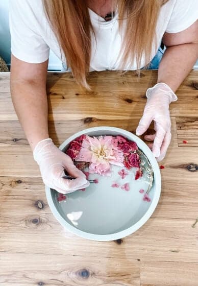 En person med langt hår arrangerer rosa og røde tørkede blomster i en rund, hvit innrammet beholder fylt med Glassklar Epoxy - CCR Epoxy Entropy SuperSap. Iført hvite hansker og hvit skjorte jobber de flittig på et trebord som minner om de som brukes til å lage elvebord. Fokuset er på hendene og beholderen.