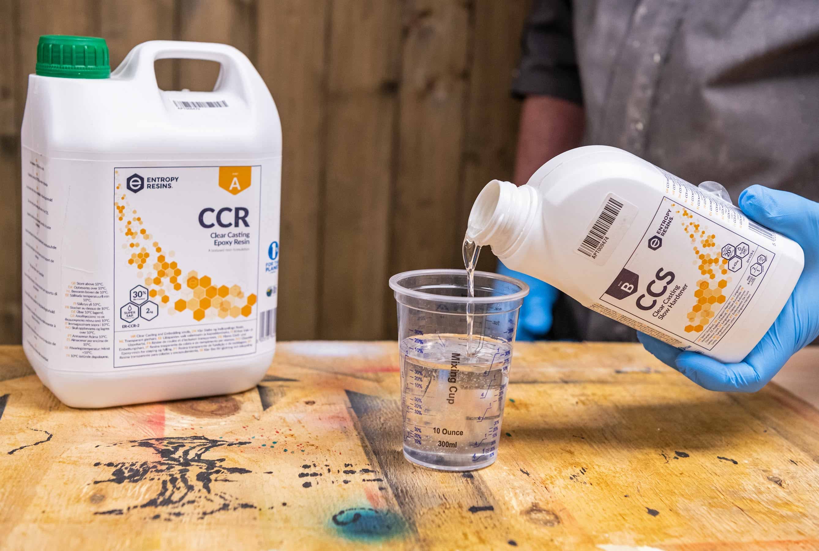 En person som har på seg blå hansker, heller klar Glassklar Epoxy - CCR Epoxy Entropy SuperSap harpiks fra en hvit flaske merket "CCR" i et målebeger på et trebord. Ved siden av koppen står en større hvit beholder med samme etikett. Bakgrunnen har treplanker som antyder elvebordprosjektet deres.