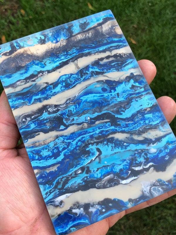 Et nærbilde av en hånd som holder et rektangulært kunstverk laget med Glassklar Epoxy - CCR Epoxy Entropy SuperSap. Kunstverket har virvlende mønstre i levende blå, hvit og svart, som minner om havbølger eller marmorert stein. Bakgrunnen består av grønt gress, litt uskarpt.