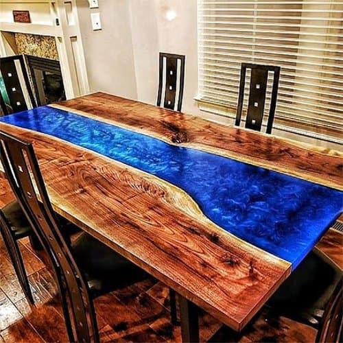 Et spisebord med en unik design har en blå epoksyharpiks-elv, laget med Glassklar Epoxy - CCR Epoxy Entropy SuperSap, som renner nedover i midten, flankert av polerte trekanter. Bordet er omgitt av seks moderne stoler med sorte polstrede seter og rektangulære utskjæringer i ryggstøttene. Persienner dekker vinduet bak bordet.