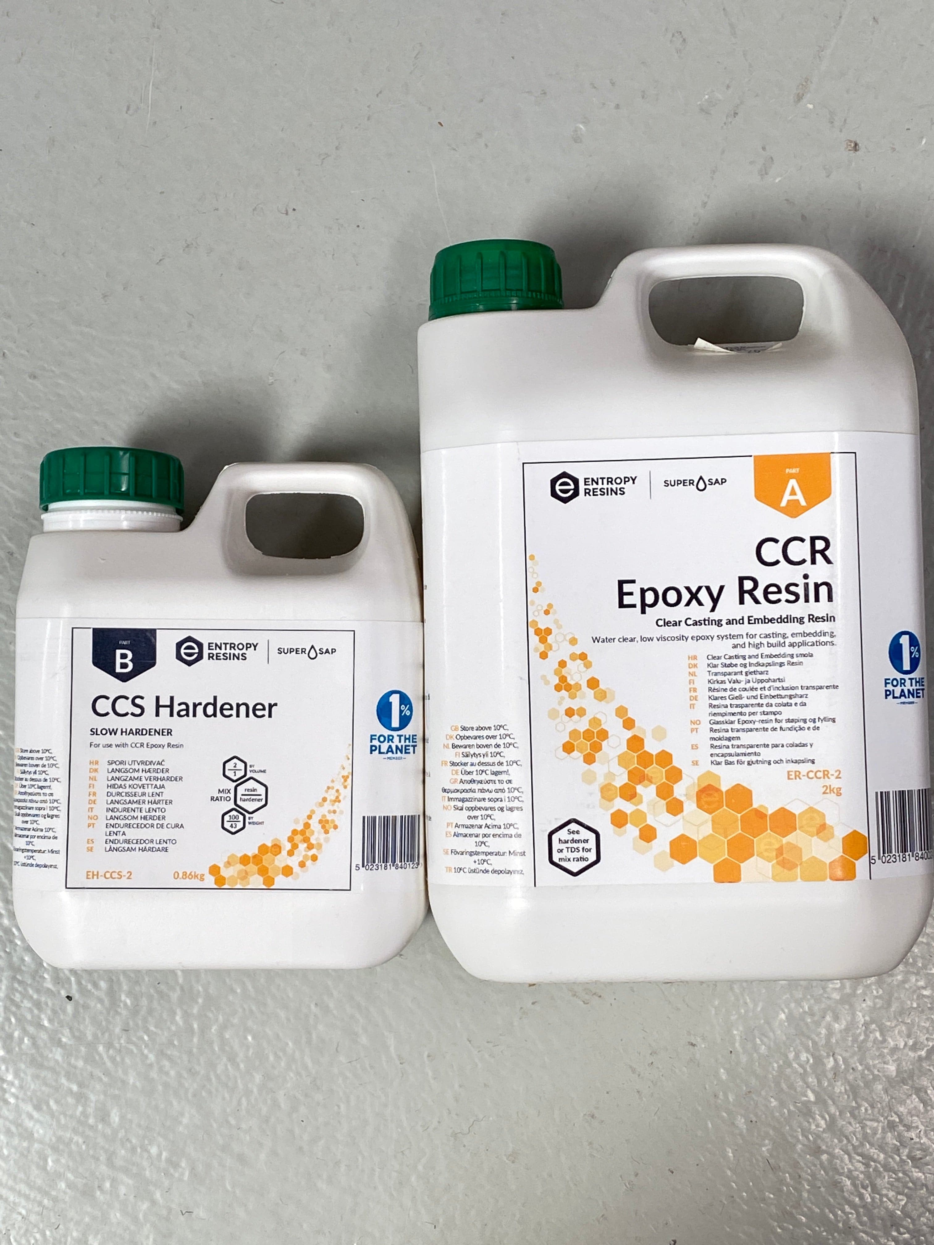 To plastbeholdere med etiketter står på en lysegrå overflate. Den større beholderen, merket "Glassklar Epoxy - CCR Epoxy Entropy SuperSap," har en grønn hette og en honeycomb-design. Den mindre beholderen, "CCS Hardener", også med en grønn hette, er til venstre, med en lignende honeycomb-design og tekst. Perfekt for å lage ditt neste elvebordmesterverk.